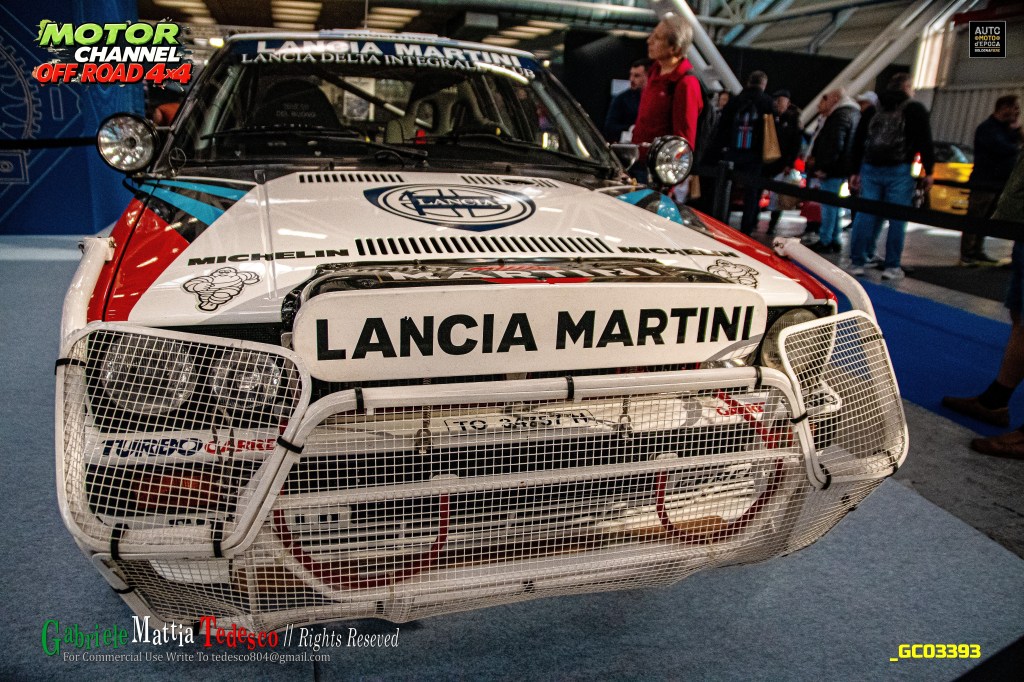 Primo piano di una Lancia Martini da rally con una griglia protettiva sul paraurti, esposta in un evento automobilistico.